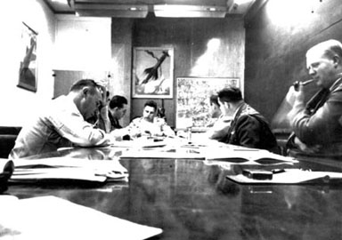 Le groupe du projet Sign dans la salle de conf&eacute;rence du d&eacute;partement T-2 &agrave; Wright Patterson, en 1948. A partir de la cauche et de la table on trouve : le lieutenant-colonel Malcolm Seashore, chef de l'Analyse Technique du Renseignement du Commandement du Mat&eacute;riel, une personne non-identifiable masqu&eacute;e par Seashore, le lieutenant-colonel J. J. Hausman, Howard McCoy ; [ce qu'on pense &ecirc;tre] le capitaine Robert Sneider, Officier de Projet sous McCoy et Clingerman, [ce qu'on pense &ecirc;tre] le colonel William Clingerman, officier executif pour l'Analyse du Renseignement du Commandement du Mat&eacute;riel (MCIA) et John "Red" Honaker, liaison avec Twining, tenant sa pipe