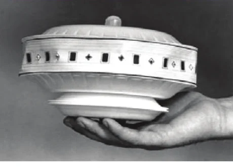 Une main tenant une maquette en forme de soucoupe faite d'assiettes, avec une bande de hublots carrés et un petit dôme