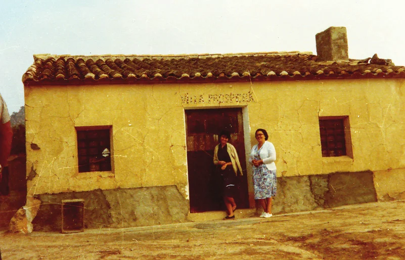 Two women at the door of a small whitewashed country house with a tiled roof, the words Villa Próspera above the door