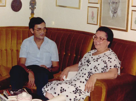 A man with glasses and a woman in a polka-dot dress sitting on a sofa in a living room
