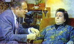 Television still of a man in a suit holding the hand of a woman sitting in an armchair with her eyes closed